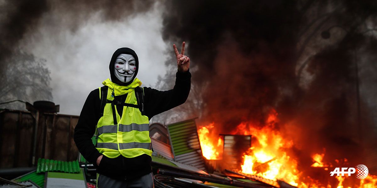 FRANCIA-SOCIAL-POLITICS-DEMO-FUEL Un manifestante que lleva una máscara de Guy Fawkes hace el signo de la victoria cerca de una barricada en llamas durante una protesta de chalecos amarillos (Gilets jaunes) contra el aumento del precio del petróleo y los costos de vida, el 1 de diciembre de 2018 en París. Abdulmonam EASSA / AFP